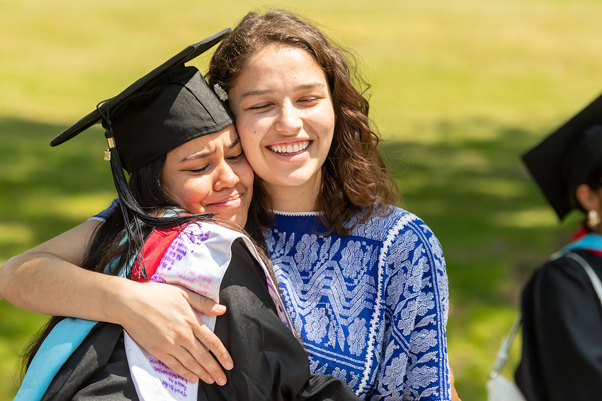 Two women embrace and smile, one of them wearing a graduation gown.